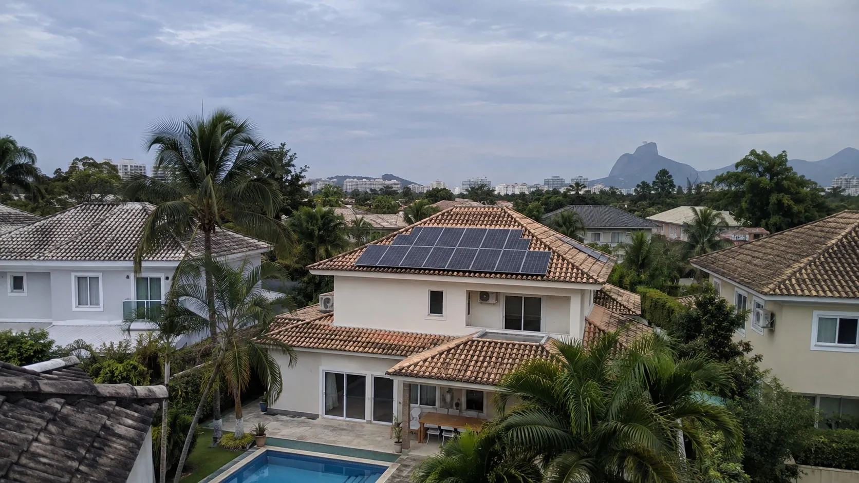 Casas de alto padrão com painéis solares, piscina e coqueiros na Barra da Tijuca, com a Pedra da Gávea ao fundo.