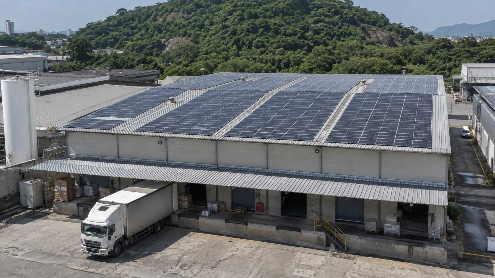 Galpão industrial com placas solares no telhado metálico, caminhão na doca e morro verde ao fundo, em Campo Grande.